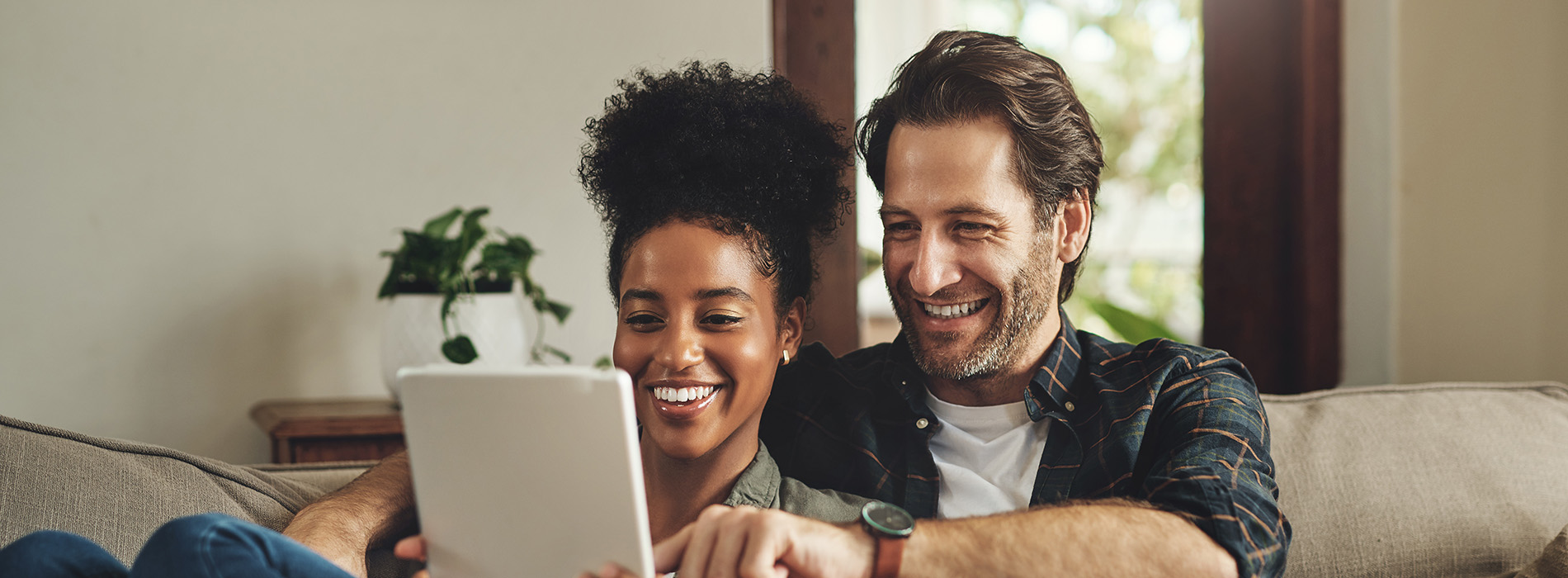 A man and woman are sitting on a couch, looking at a tablet with smiles on their faces.