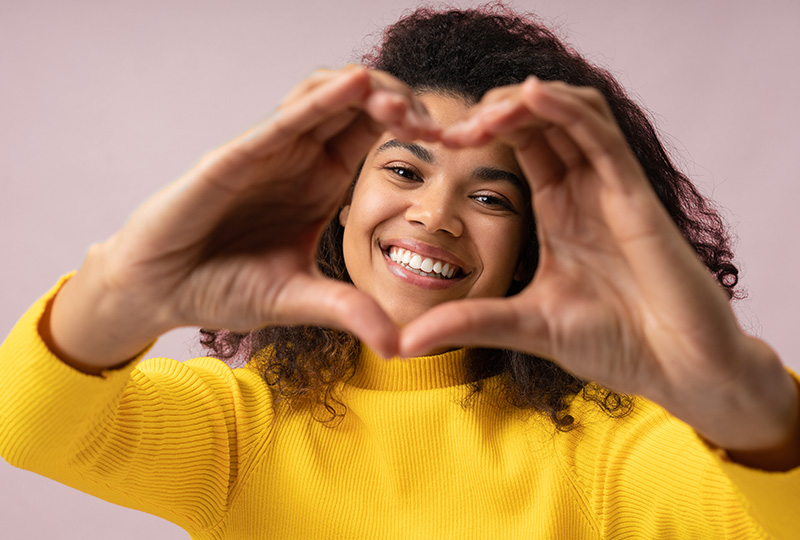 A woman with dark hair wearing a yellow top holds her hands together in a heart shape.