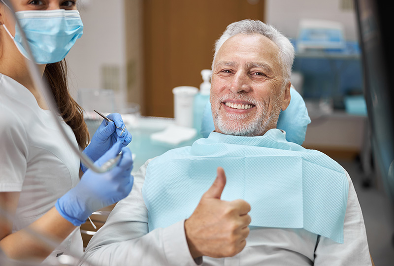 The image features an older man with a beard sitting in a dental chair, giving a thumbs-up gesture, surrounded by dental professionals in a dental office setting.