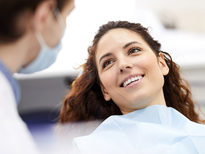 This is a photograph featuring a smiling woman seated in a dental chair with a dentist standing beside her, both wearing medical masks. The setting appears to be a dental office, and they seem to be engaged in a professional interaction.