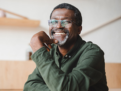The image features a man with glasses sitting at a table, smiling slightly and leaning on his hand, with a blurred background that appears to be an indoor setting.