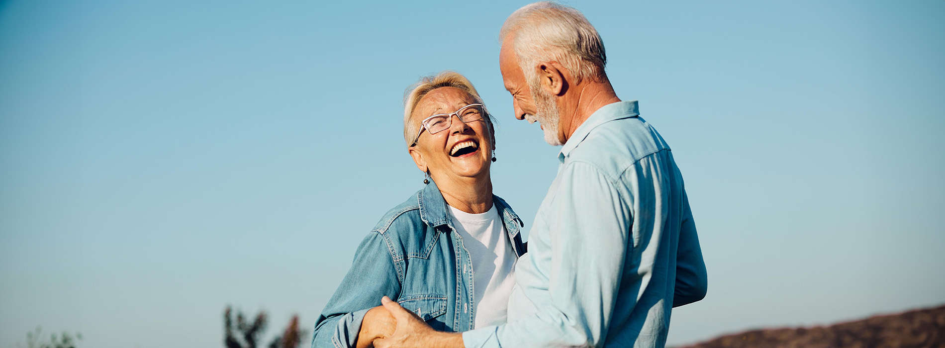The image shows two elderly individuals, presumably a man and woman, outdoors during daylight, smiling at each other with one arm around the other s waist, both dressed casually, standing on what appears to be a grassy area under a clear sky.