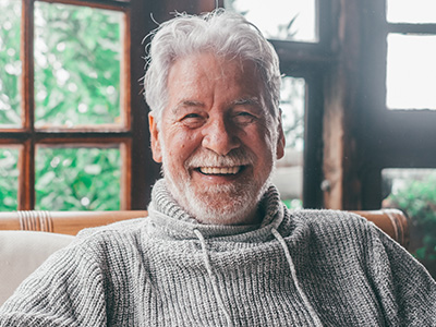 The image features an elderly man with white hair, wearing a sweater, sitting indoors with a smile on his face.