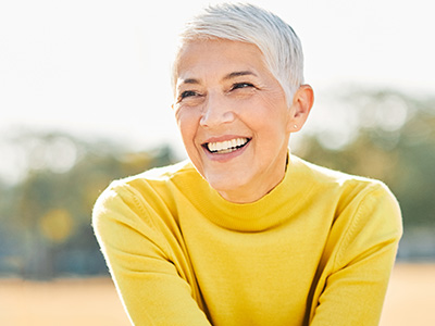 A smiling older woman with short hair wearing a yellow top, sitting outdoors against a blurred background.
