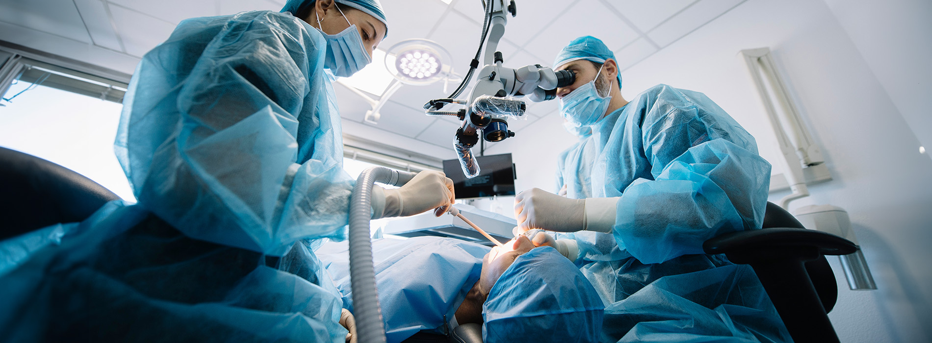 The image shows two medical professionals wearing surgical gowns and masks, with one person operating a microscope while the other appears to be assisting or observing during what might be a medical procedure.