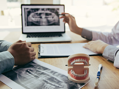 Two people sitting at table with laptop, model teeth, and medical images displayed on screen.