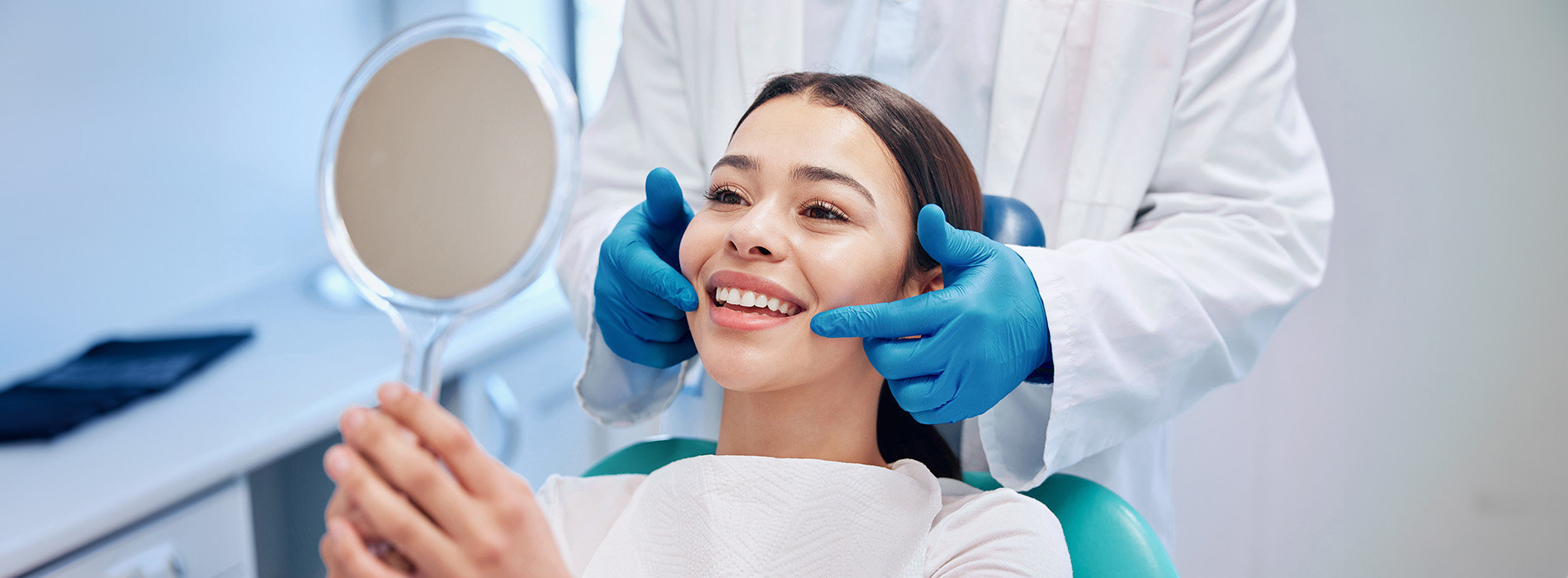 The image shows a dental professional performing an examination on a patient s teeth while the patient smiles, with both individuals wearing face masks and the professional using a mirror for inspection.