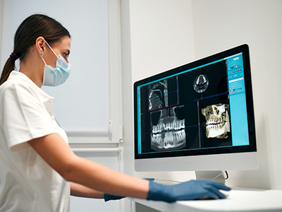 Woman wearing a white lab coat, blue gloves, and a face mask operating a medical imaging machine with a large screen displaying an X-ray.