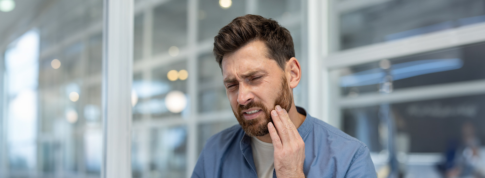 A man with a beard is captured from behind, touching his face with both hands while standing indoors.