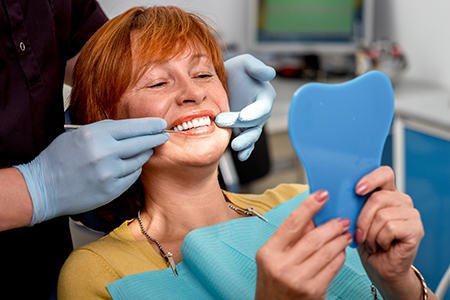 A woman sitting in a dental chair with a blue facial mask, smiling at the camera while holding up a toothbrush.
